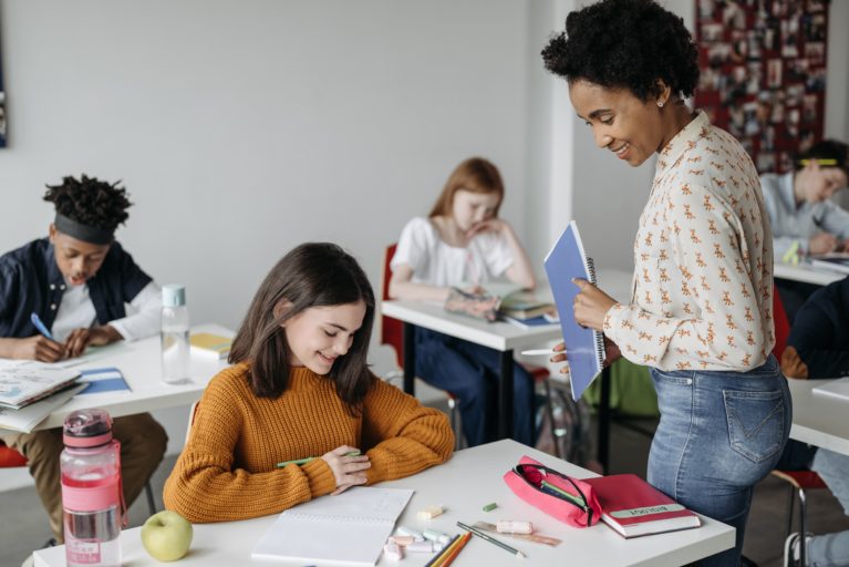 Na imagem estão presentes jovens estudando em sala de aula, e, ao lado da carteira de uma aluna, a professora a orienta. 