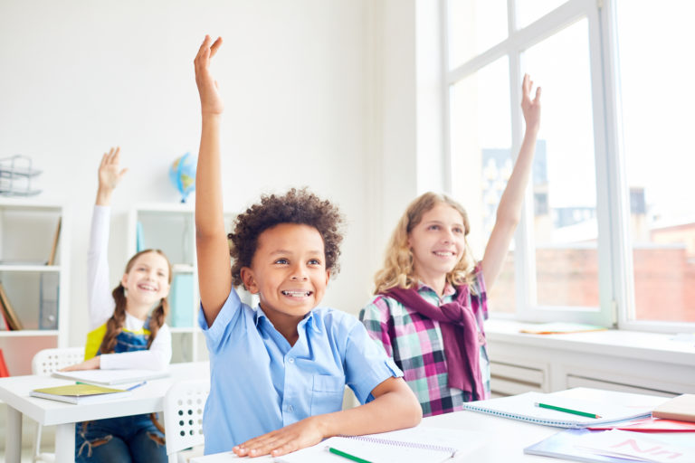 Imagem de três crianças em uma sala de aula sentadas em frente a mesas escolares. Todos estão com os braços levantados e sorrindo.