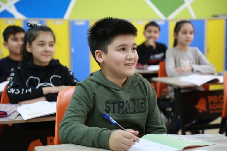 Um grupo de alunos aparece em uma sala de aula, olhando para a frente e há um garoto de blusa verde em destaque, com uma caneta na mão.