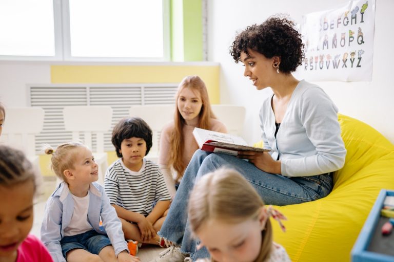 Professora de educação infantil está sentada em um puff amarelo e lê uma história para alguns alunos espalhados.
