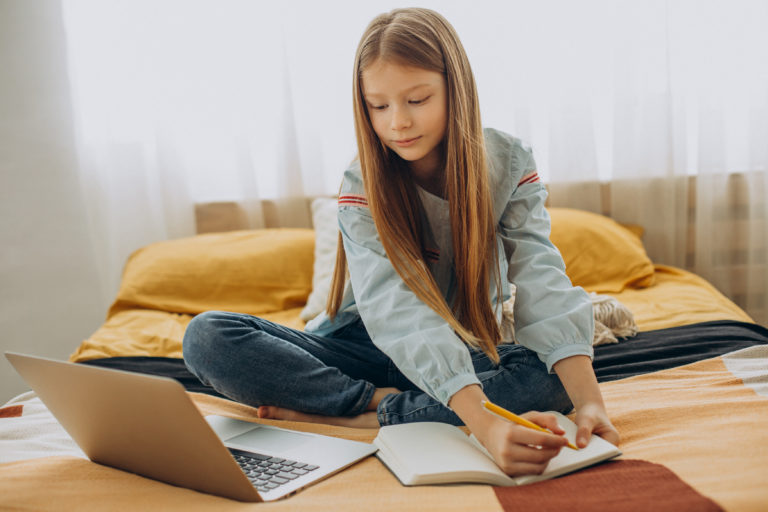 Imagem de uma menina sentada em uma cama enquanto se dedica a melhorar seu desempenho escolar dentro de um quarto. Ela está olhando para um notebook enquanto faz anotações em uma agenda.