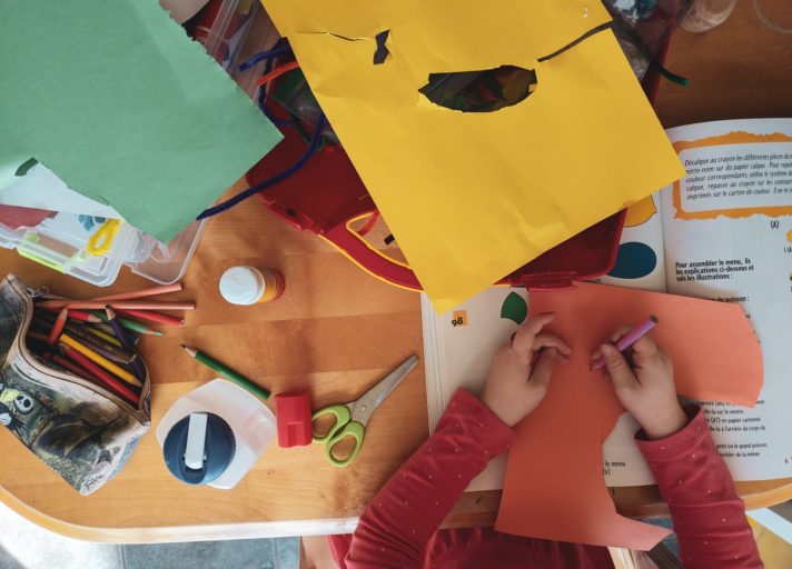 Visão de cima de uma mesa com materiais escolares, como cartolinas coloridas, lápis de cor, tesoura, cola, livro e temos uma criança mexendo.
