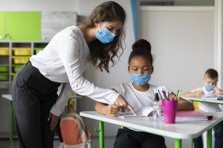 Imagem de uma professora e uma aluna em uma sala de aula. A aluna está sentada em frente a uma mesa enquanto a professora a ajuda com a tarefa. Ambas usam máscara.