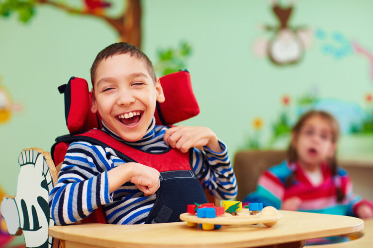 Foto de um garoto com paralisia cerebral na escola, sentado e sorrindo brincando com um objeto educativo de madeira.