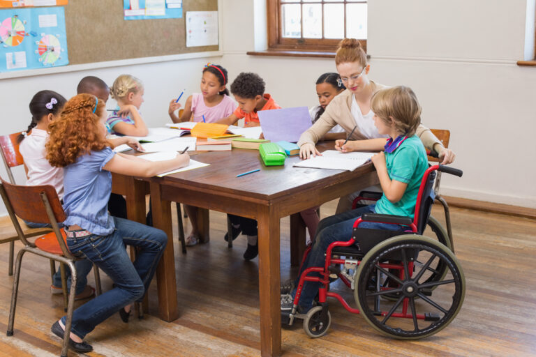 A foto de uma sala de aula, com estudantes e professora em volta de uma mesa e há um garoto em uma cadeira de rodas, representando a inclusão social na escola.