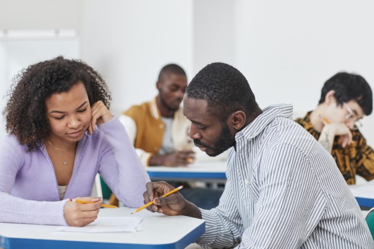 Em uma sala de aula, observamos um professor negro, ajudando uma estudante negra a realizar uma avaliação somativa. No fundo da imagem, há outros estudantes também realizando a prova.