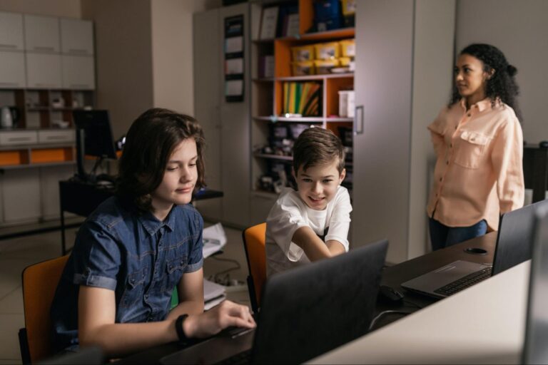 Dois estudantes em uma sala de aula de informática utilizando computadores com a supervisão de uma professora. 