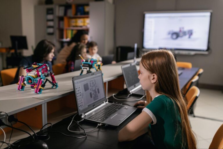 Imagem de uma garota ruiva utilizando um notebook em uma sala de informática; ao fundo, desfocados, estão mais quatro estudantes usando outro computador.