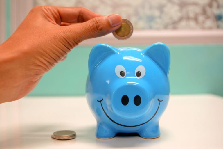 Uma pessoa está colocando uma moeda em um cofrinho em formato de porco na cor azul que está em cima de uma mesa. 