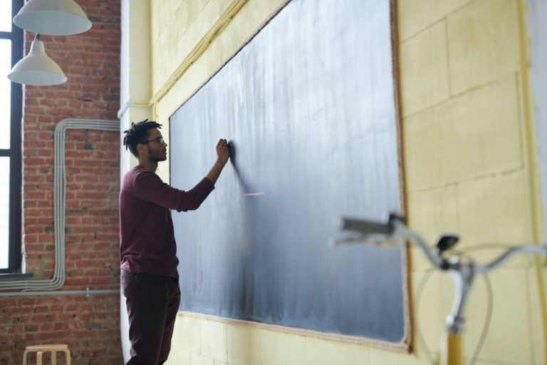 Homem negro, jovem, escreve em um quadro negro com um giz. Ele se encontra em uma sala de aula. 