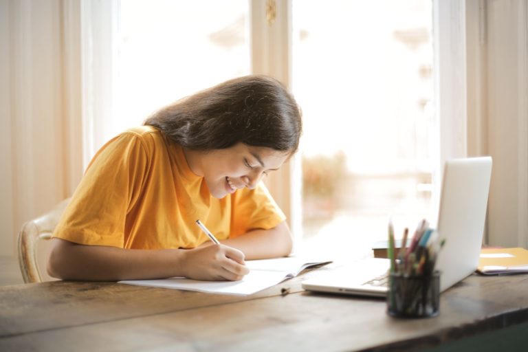 Garota de camiseta amarela escreve em seu caderno, em frente a um notebook aberto.