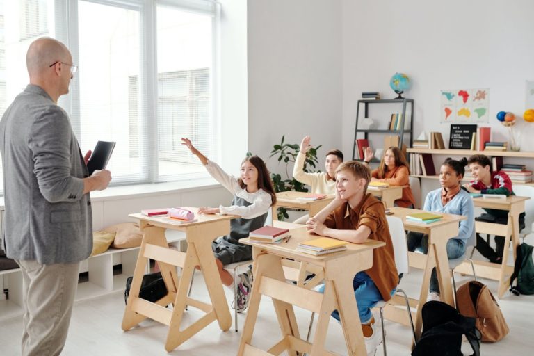 A imagem mostra uma sala de aula com uma turma de seis alunos, interagindo com o professor que está logo à frente das crianças. 
