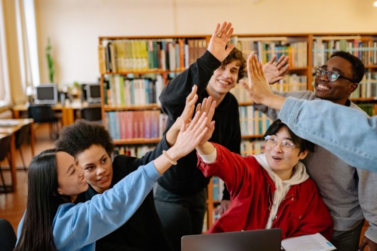 A imagem mostra um grupo de cinco jovens em uma biblioteca, eles estão em frente a uma notebook aberto e batem as mãos umas nas outras em comemoração.