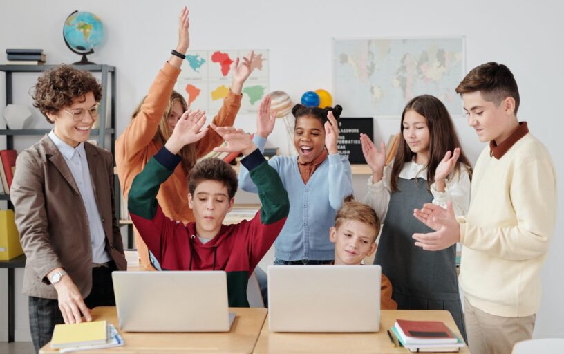 Ao lado de uma professora os alunos da esquerda comemoram a vitória e os alunos da direita estão batendo palmas para equipe vencedora, ambos estão em frente a computadores.
