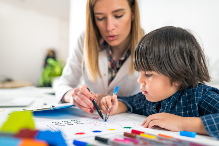 Psicóloga branca, loira de cabelos lisos, segurando uma caneta preta apontando para o papel que a criança, seu paciente, está desenhando.