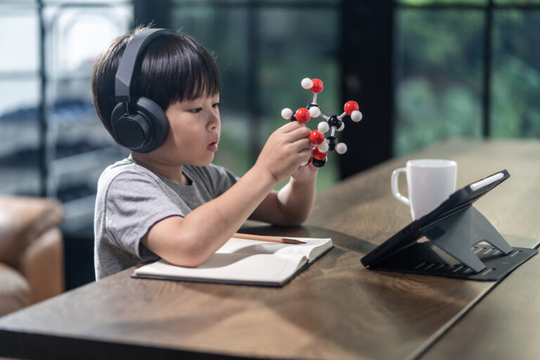 Um garoto asiático está sentado em frente a uma mesa, com fones de ouvido, um tablet e um caderno e desenvolvendo o raciocínio lógico com um modelo de células de metal.