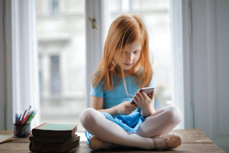 Criança branca, ruiva de cabelos curtos e lisos, sentada em cima de uma mesa de madeira, com um smartphone em mãos.