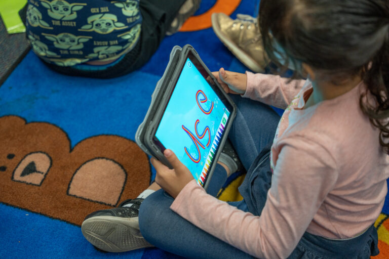 1st grade student uses technology in the classroom by practicing writing the sight word "use." She uses a rainbow drawing tool on her iPad.