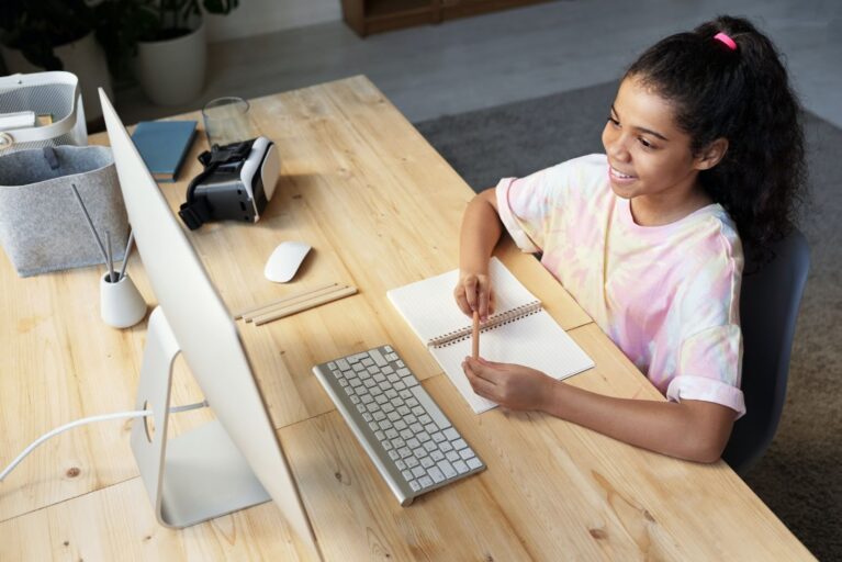 A imagem mostra uma garota estudando em frente a um computador que está sob uma mesa de madeira. 