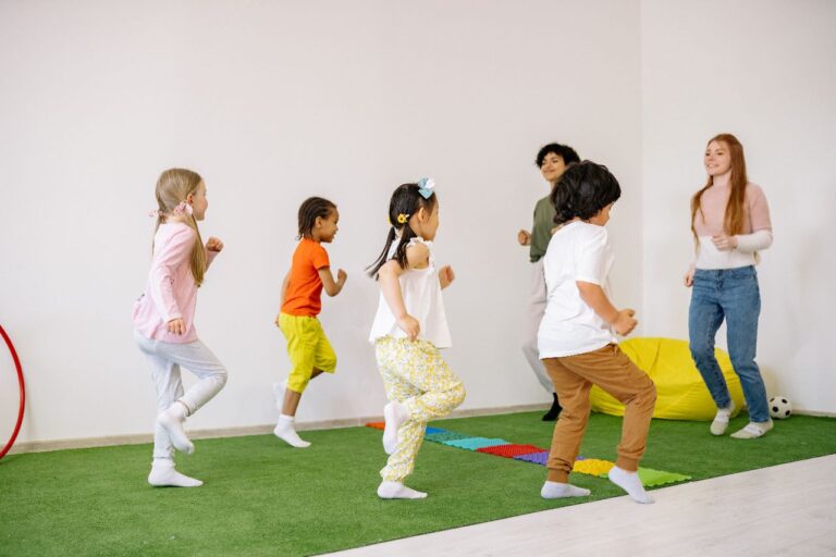 Em uma sala de aula lúdica, com tapete que imita grama, há quatro crianças brincando, com duas professoras à frente.