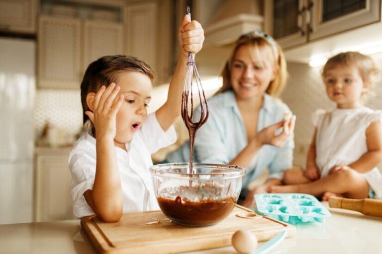 Uma família composta por uma mãe e dois filhos estão cozinhando. Enquanto o menino faz uma mistura de chocolate em uma vasilha, a mãe junto a filha observam a criança.