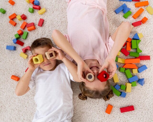  Duas crianças estão deitadas no chão, com peças de brinquedo ao redor, enquanto sorriem. Elas estão segurando duas peças em cima de seus olhos.
