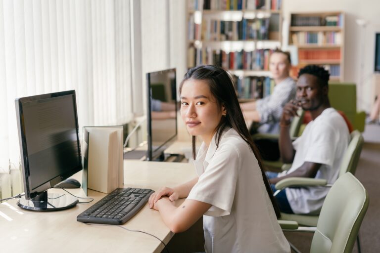 Imagem de uma sala de informática, com computadores, teclados e cadeiras para os alunos. Em foco, há uma mulher com traços asiáticos olhando para a câmera, ela possui cabelos pretos e usa uma camisa branca. Ao fundo, desfocados, vemos dois homens também olhando para a câmera. 