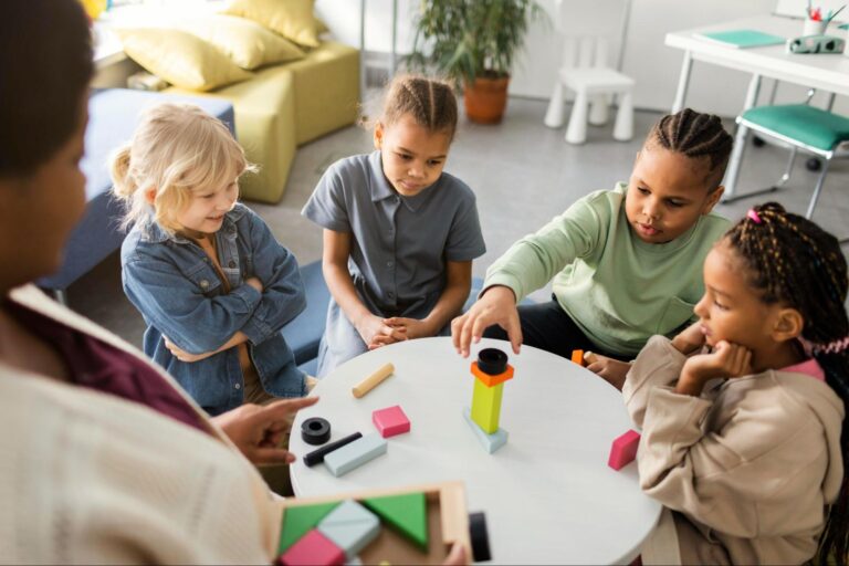 Quatro crianças brincando com peças de montar estão sentadas em volta de uma mesa, enquanto a professora, que está em pé, instrui a aprendizagem.