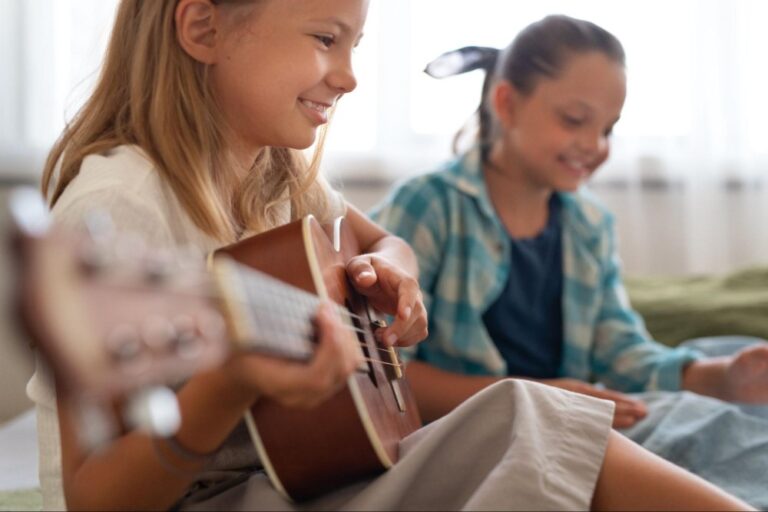 Duas meninas sentadas no chão estão sorrindo, enquanto uma delas toca violão. 