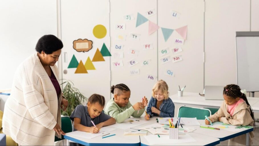 Quatro crianças estão desenhando em alguns papéis em volta de uma mesa com a professora ao lado, instruindo a aprendizagem. 