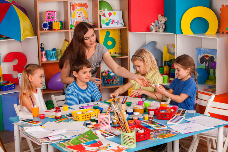 Em uma sala de aula de arte na educação infantil, uma professora conversa com quatro crianças que estão em volta de uma mesa cheia de materiais de pintura.