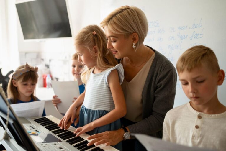 Mulher sentada em um banco, com crianças ao redor e uma em sua frente, está ensinando partituras de teclado durante uma aula de música.