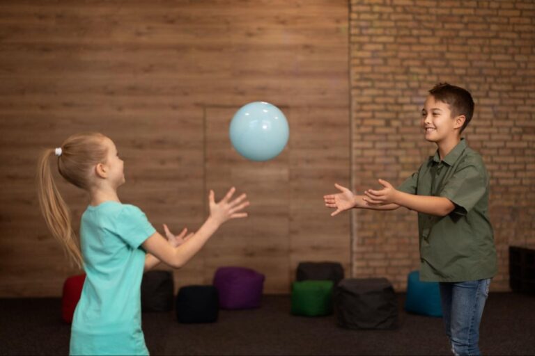 Duas crianças brincando com uma bola azul. Ao fundo, existem alguns puffs coloridos espalhados.