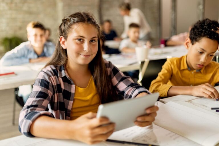Uma menina sorridente e vestida com camiseta rosa e camisa xadrez, está sentada em uma cadeira na sala de aula enquanto segura um tablet nas mãos. Ao redor, vemos outras crianças estudando.