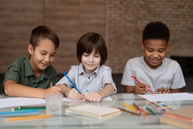 Três crianças estão sorrindo enquanto olham para o papel em uma mesa, cada um segura um lápis em cores diferentes. Também há lápis e caderno espalhados na superfície. 