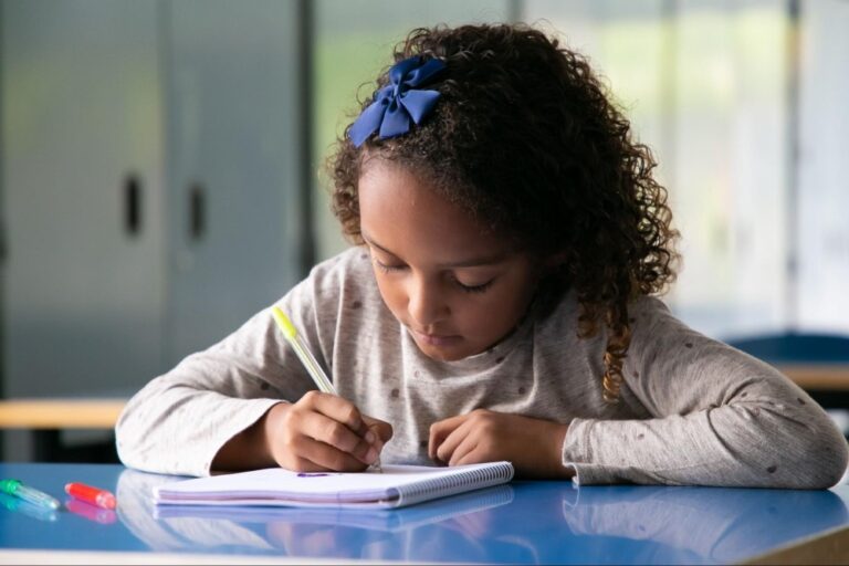Criança de cabelos cacheados e um lacinho azul está em uma sala de aula, em sua mão há uma caneta amarela que está usando para escrever em um caderno. Na mesa azul, há alguns outros tipos de canetas.