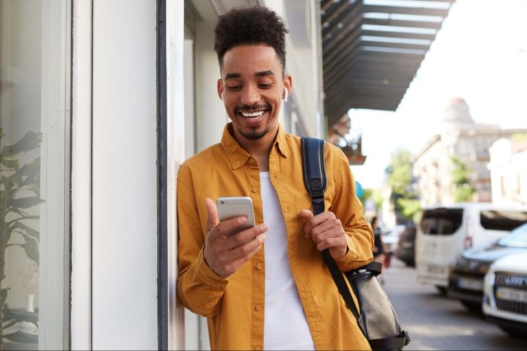 Homem com camisa amarela e mochila no ombro está encostado em uma parede enquanto segura seu celular na mão e sorri. Ao fundo vemos carros estacionados e a calçada.