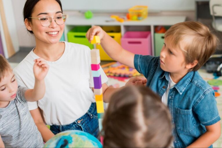 Na imagem, há uma mulher com três crianças ao lado, que estão montando uma torre colorida. Ao fundo, há algumas caixas coloridas. A imagem representa a afetividade em sala de aula.