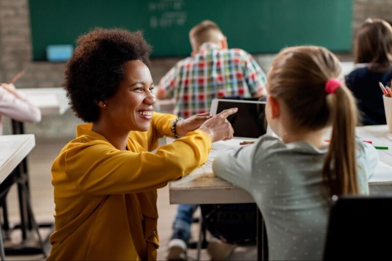 Professora ao lado de aluna sentada à mesa escolar orientando no uso de celular em sala de aula