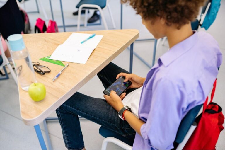 Criança sentada em carteira escolar fazendo uso de celular na sala de aula.