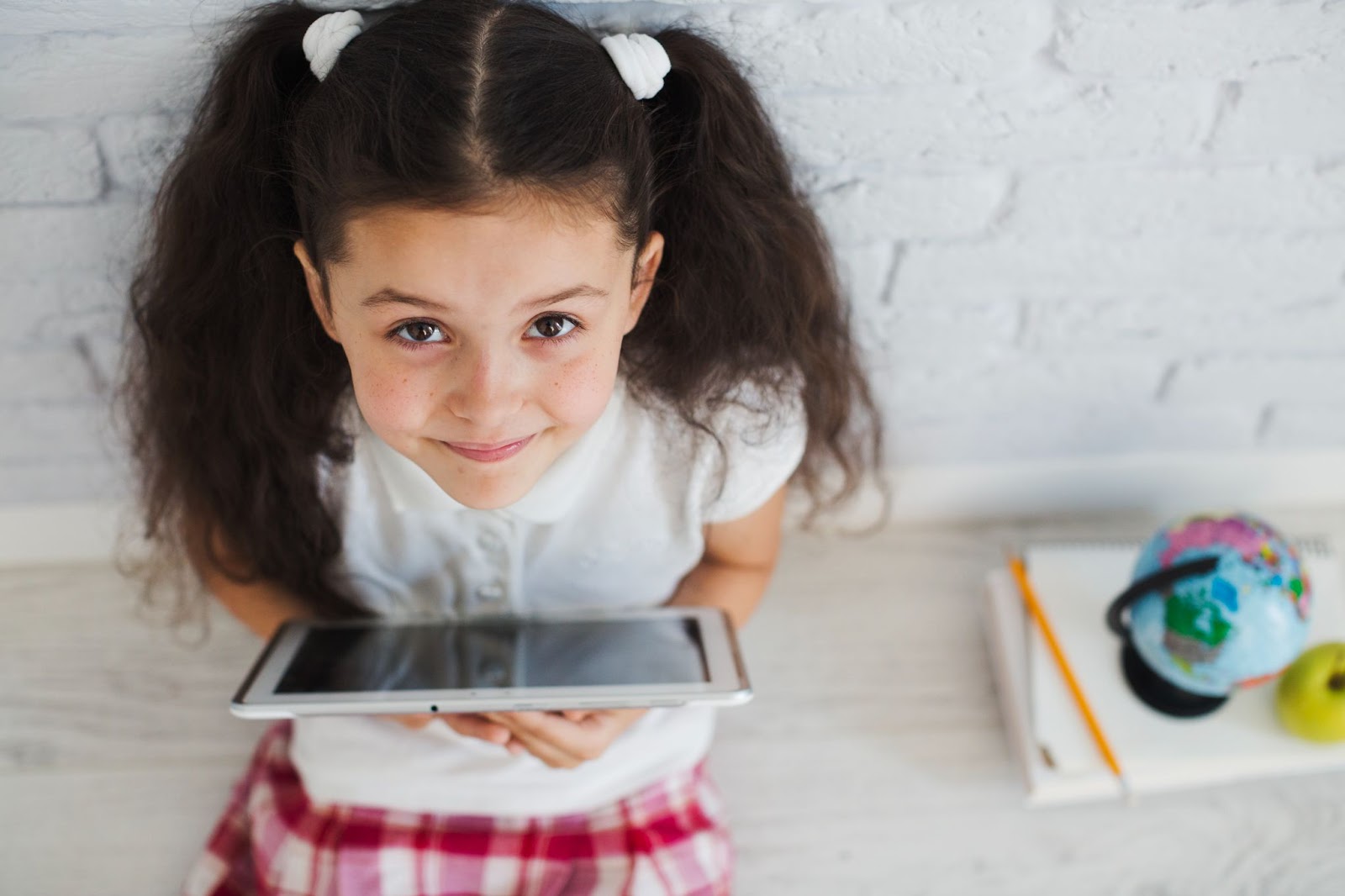 Uma menina sentada em um banco de madeira segura um tablet enquanto olha para cima. Ao seu lado há um caderno, um lápis e um pequeno globo terrestre.