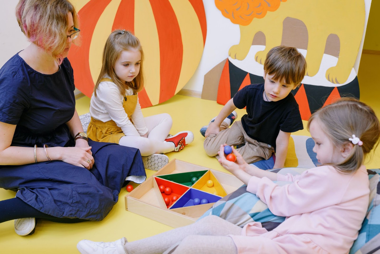 Na imagem uma professora de azul ensina três crianças formas geométricas educação infantil com objetos no formato de quadrado, triângulo e círculo.