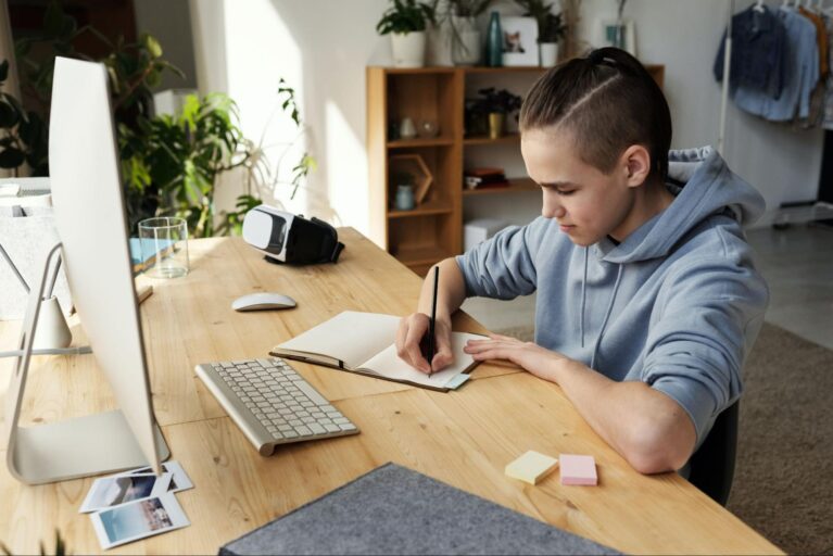 Um menino está sentado na frente de uma escrivaninha, no computador, anotando algo em um caderno.