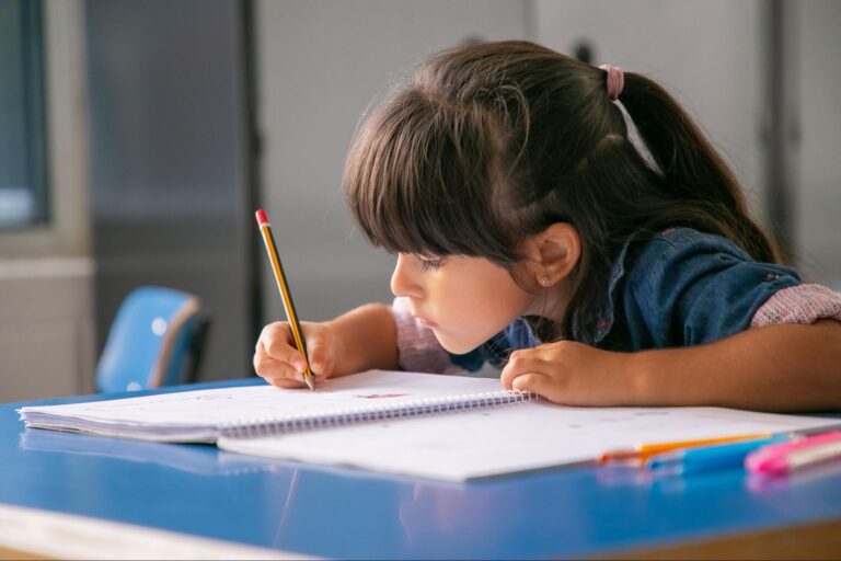 Criança segurando um lápis para fazer as atividades no caderno. Em sua mesa, há algumas canetas. 