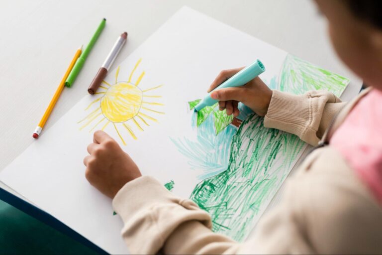 Criança está desenhando em uma folha branca, no desenho há uma árvore na grama verde e um sol. A criança está segurando um lápis azul para pintar o céu.