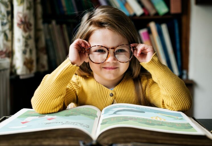 Na imagem, há uma criança de óculos, sentada em frente a um livro ilustrado e com poucas linhas escritas. Ao fundo, há uma estante com diversos livros em diferentes cores.