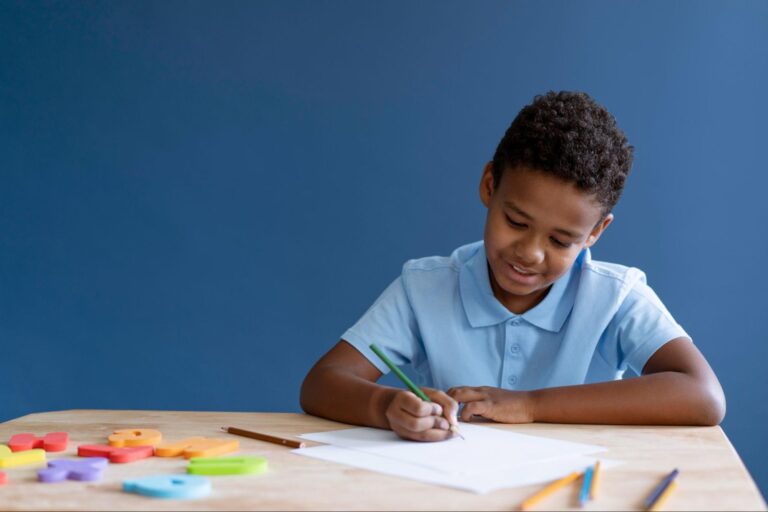 Criança sorrindo enquanto segura um lápis verde em sua mão. Na mesa, há algumas letras coloridas e diferentes cores de lápis.