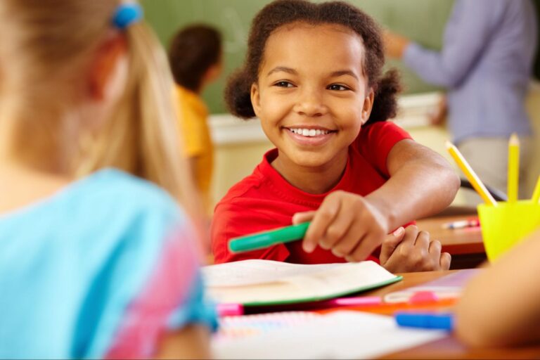 Na imagem há uma criança sorrindo enquanto entrega uma caneta verde para outra criança em sua frente, o ambiente é uma sala de aula.