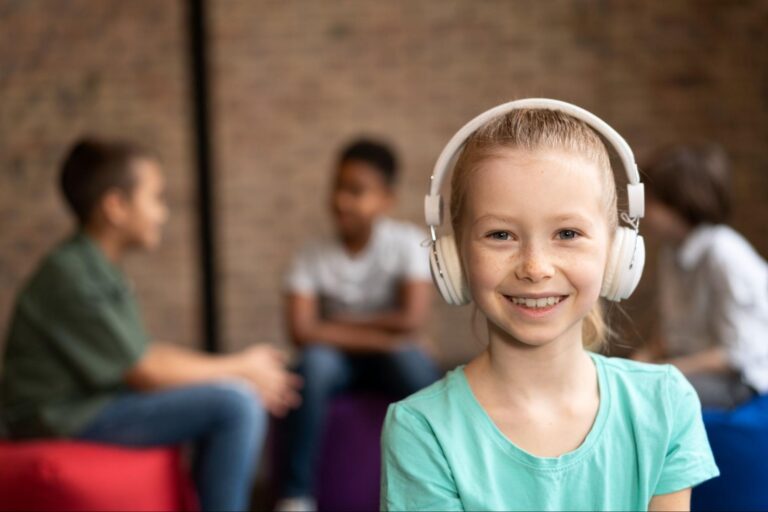 Menina de blusa verde-água está sorrindo com um fone em sua cabeça. Ao fundo, existem outras crianças sentadas e conversando entre si.