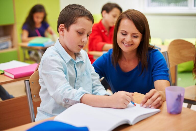 Na imagem há uma professora abaixada ao lado de um aluno, que está escrevendo em seu caderno em uma sala de aula.